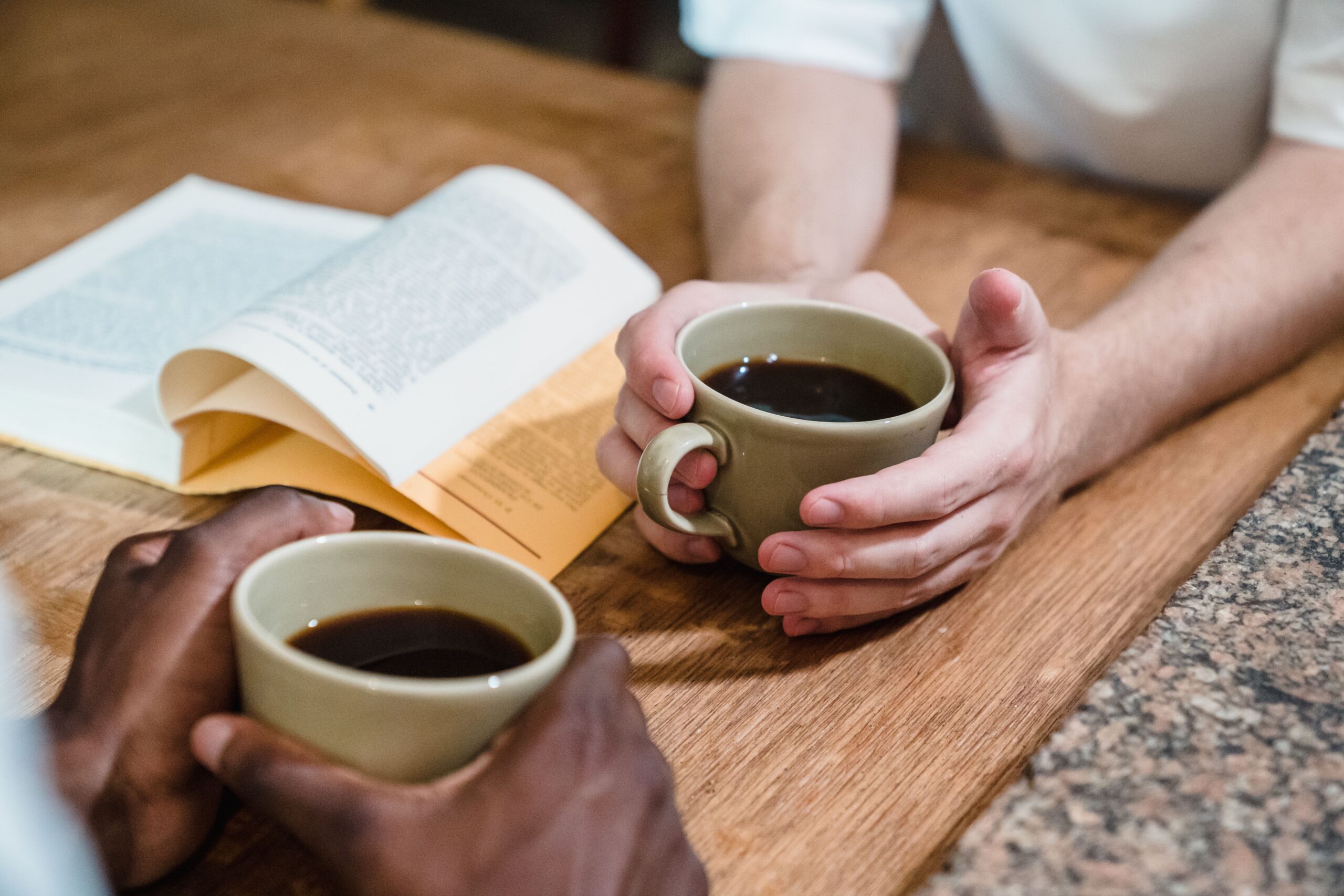 Two people holding coffee cups and reading a book on a wooden table.